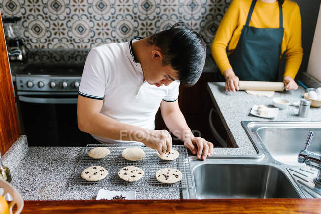 High angle of Latin teenage boy with Down syndrome decorating raw cookies with chocolate chips while cooking in kitchen at home — Stock Photo