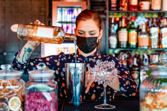 Camarera en máscara protectora vertiendo bebida alcohólica en la coctelera mientras prepara cóctel y trabaja en el bar durante el coronavirus - foto de stock