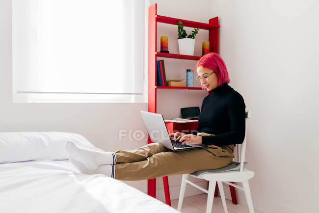 Vista lateral de una joven freelancer enfocada en ropa casual sentada en una silla y usando un portátil mientras trabaja en un proyecto en un apartamento moderno y luminoso - foto de stock