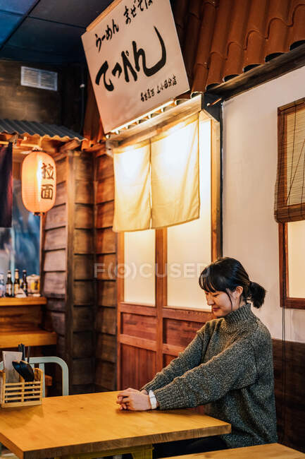 Задоволена азіатка у випадковому светрі, що дивиться вниз, сидячи за дерев'яним столом у барі. — стокове фото