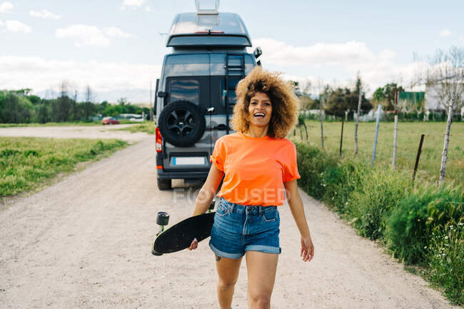 Feliz hembra afroamericana llevando longboard y mirando a la cámara con sonrisa mientras camina por el camino del campo cerca de la furgoneta en verano - foto de stock