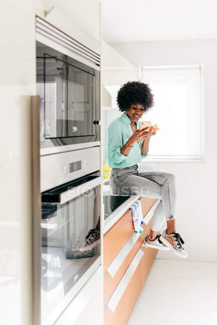 Moderne schöne Afroamerikanerin mit Smartphone in der Hand sitzt auf dem Küchentisch und schaut zu Hause weg und isst Apfel — Stockfoto