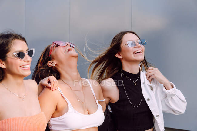Amigas positivas en gafas de sol de moda y ropa casual riéndose felizmente mientras pasan un rato agradable juntas - foto de stock