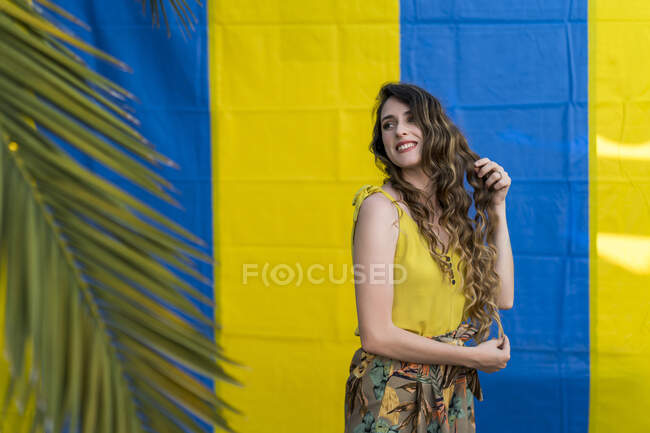 Deleitado hembra tocando el pelo largo ondulado mientras está de pie sobre dos fondo de color en el parque con palmera - foto de stock