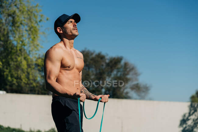 Forte atleta maschio con busto nudo che si esercita con elastico sul prato alla luce del sole — Foto stock