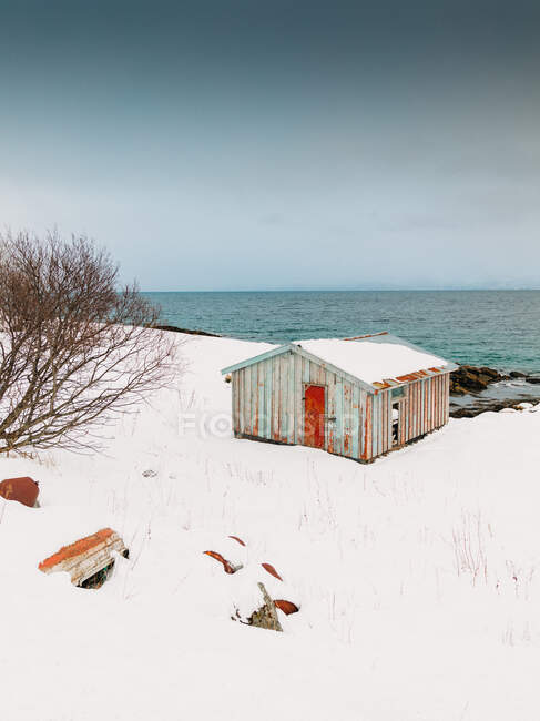 Cabana de madeira desgastada localizada na costa branca de neve do mar contra o céu azul sem nuvens nas ilhas Lofoten, Noruega — Fotografia de Stock