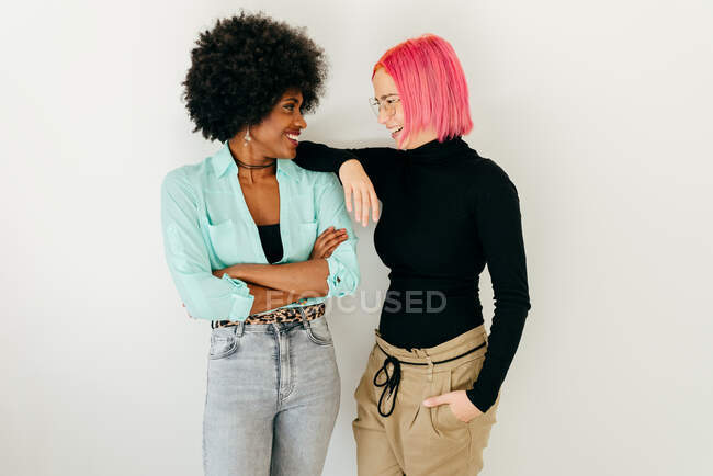 Cheerful young pink haired woman and African American girlfriend in stylish outfit standing looking at each other on white background — Stock Photo