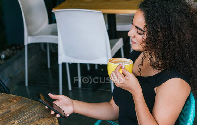 Vista lateral de la joven mujer hispana feliz navegando por el teléfono móvil mientras disfruta de un delicioso capuchino en la mesa de la cafetería de la calle - foto de stock