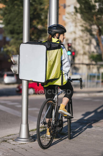 Back view of delivery woman with blank thermal bag looking away and sitting on bicycle on pavement near road in city — Stock Photo