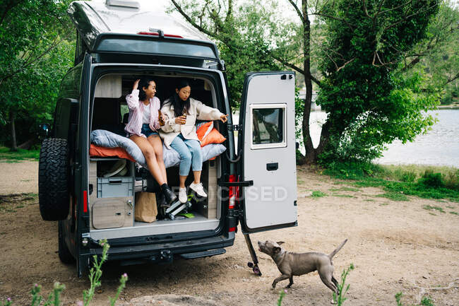Traveling young multiracial girlfriends drinking beer and giving command to dog while having fun and resting together in camper van parked near river in countryside — Stock Photo