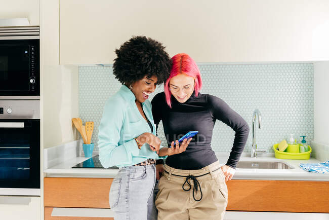 Jovens amigas multirraciais alegres em roupas da moda usando telefone celular e discutindo notícias nas mídias sociais enquanto estão na cozinha em casa — Fotografia de Stock