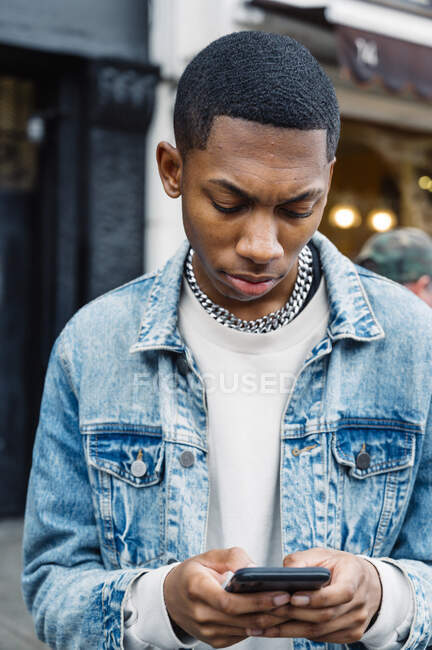 Front view of black positive young man in jeans outfit messaging on mobile phone while walking in city — Stock Photo