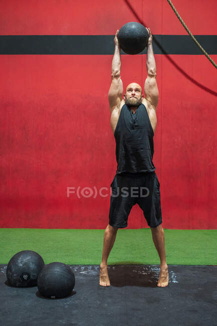 Voller Länge starker männlicher Athlet, der schweren Ball in erhobenen Armen hebt, während er in einem modernen Fitnessstudio trainiert — Stockfoto