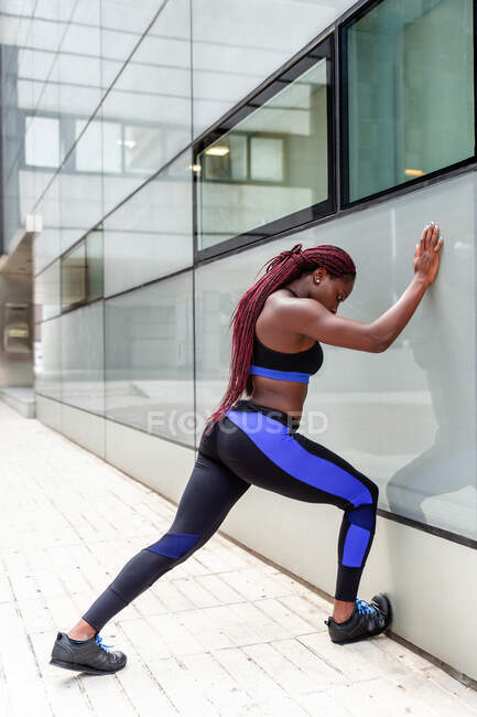 Side view of young muscular ethnic woman leaning on wall during workout on street — Stock Photo