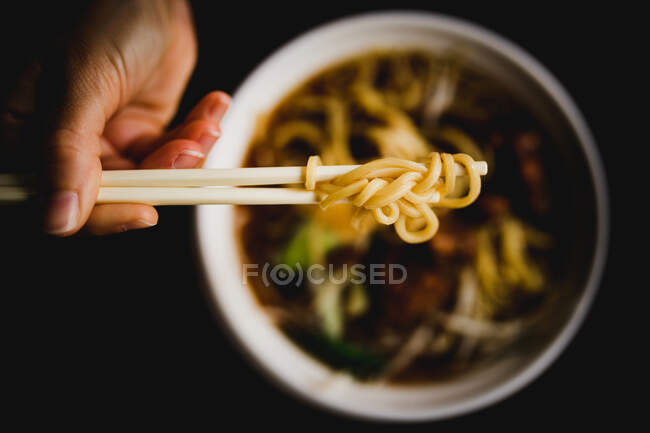 Gros plan de la main d'une personne anonyme utilisant des baguettes pour prendre des nouilles dans un bol avec une délicieuse soupe asiatique — Photo de stock