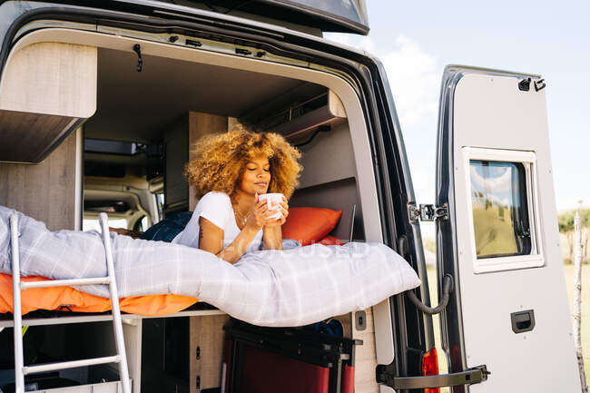 Afro-americana com cabelos encaracolados deitada na cama e desfrutando de bebida quente fresca na manhã de verão no campo — Fotografia de Stock