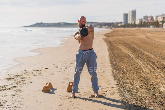 Ganzkörperglatzkopf und hemdloser Mann, der in die Kamera schaut und Schaukeln mit der Kettlebell macht, während er barfuß an der Sandküste mit der Stadt im Hintergrund steht — Stockfoto