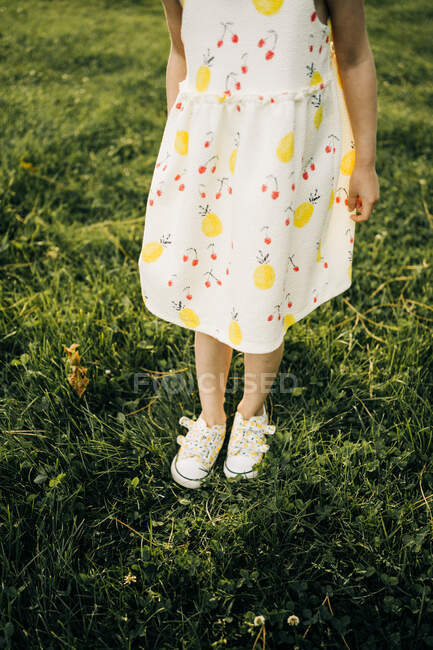 Зверху врожаю анонімна маленька дівчинка в білому сараї з квітковим візерунком і взуттям, що стоїть на зеленому трав'янистому лузі в літній день — стокове фото