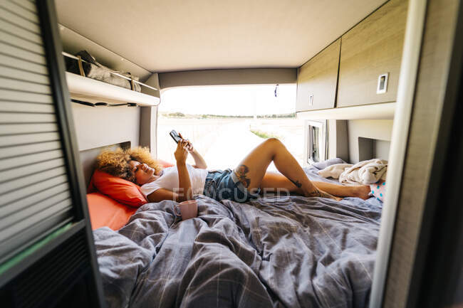 Anonymous woman with Afro hairstyle lying upside upon bed in trailer and browsing mobile phone on sunny summer day — Stock Photo