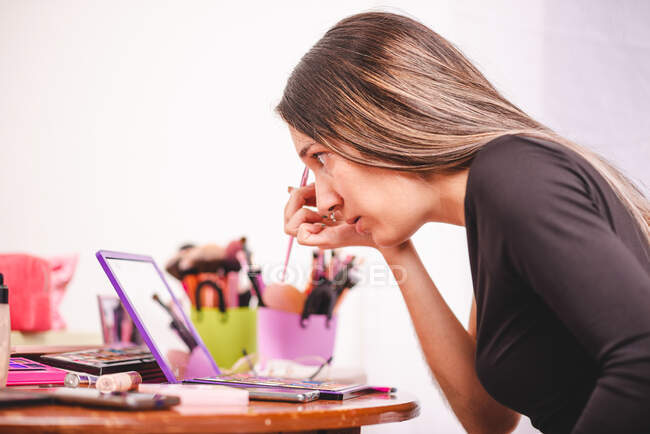 Joven mujer mirando el espejo y la pintura ornamento en la cara mientras se aplica maquillaje creativo en el estudio - foto de stock