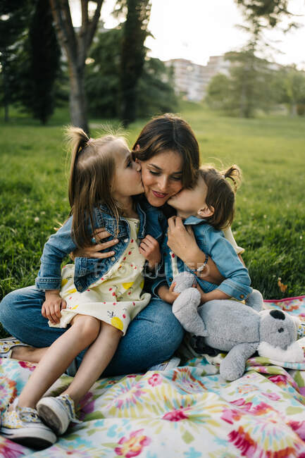 Linda hermanas pequeñas besar feliz madre mientras descansan juntos en manta en el césped verde en el parque de verano - foto de stock
