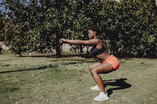 Vue latérale de la jeune femme afro-américaine qui fait des squats dans le parc par une journée ensoleillée — Photo de stock