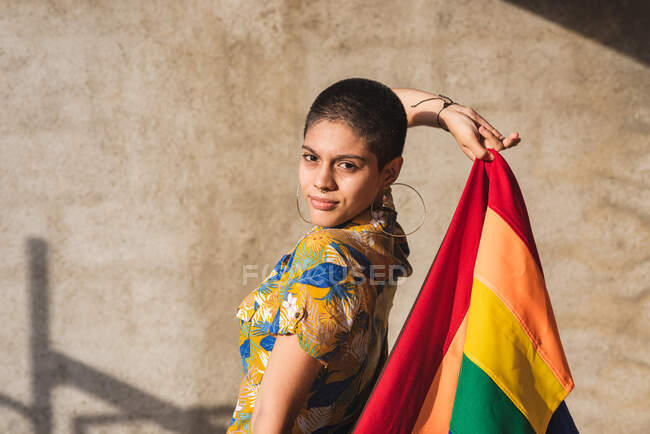 Ernsthafte junge bisexuelle ethnische Frau mit bunter Flagge, die LGBTQ-Symbole darstellt und an sonnigen Tagen in die Kamera blickt — Stockfoto