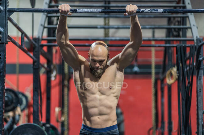 Muscular bald male with beard hanging on metal bar and doing pull ups during intense workout in gym — Stock Photo