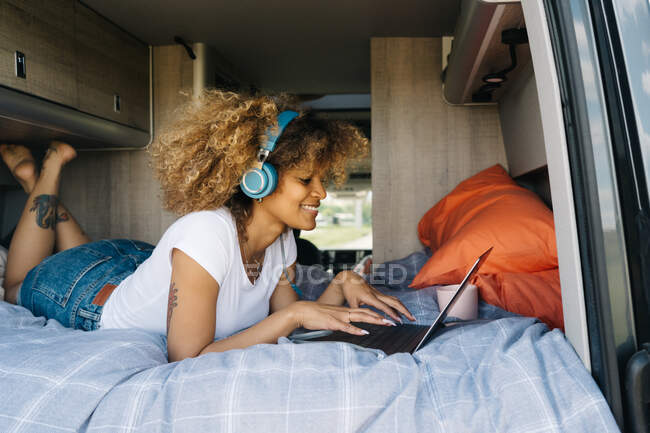Alegre mujer afroamericana sonriendo y escuchando música en auriculares mientras está acostada en la cama en caravana y navegando en las redes sociales en el portátil - foto de stock