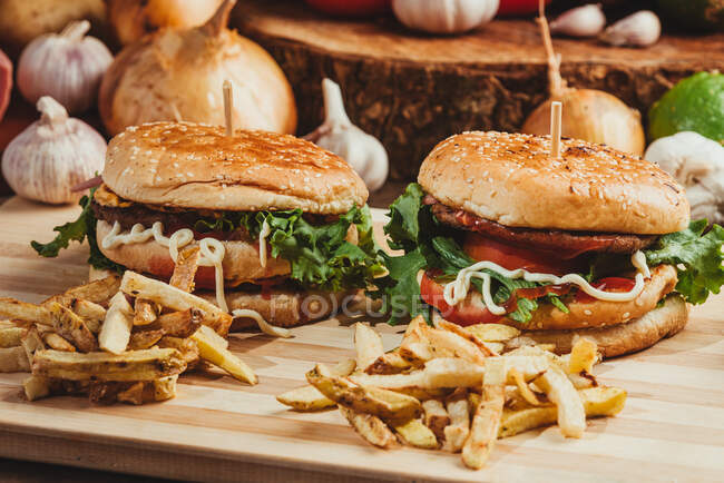 Hamburguesas apetitosas con verduras y chuletas colocadas sobre tabla de madera con papas fritas en la cocina - foto de stock