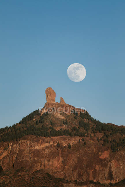 Ефектний краєвид з великим повним місяцем на блакитному небі над скелястим гірським піком з зеленим лісом в літній вечір — стокове фото