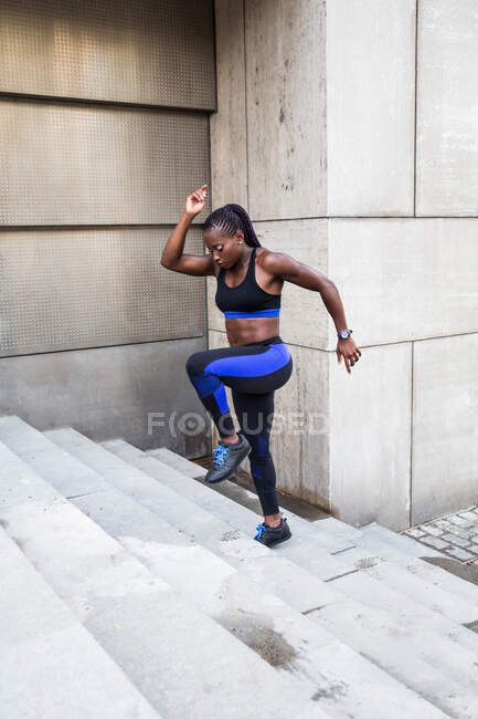 Vista lateral da mulher afro-americana forte correndo escadas enquanto treina perto do edifício moderno na rua da cidade — Fotografia de Stock