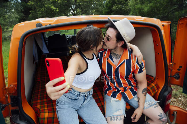 Casal encantado de viajantes sentados em van e tomando auto-tiro no smartphone enquanto abraça e desfruta de aventura de verão — Fotografia de Stock