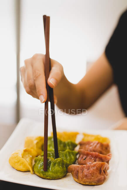 Hand of woman holding bamboo chopsticks and eating colorful dumplings from white ceramic square plate — Stock Photo