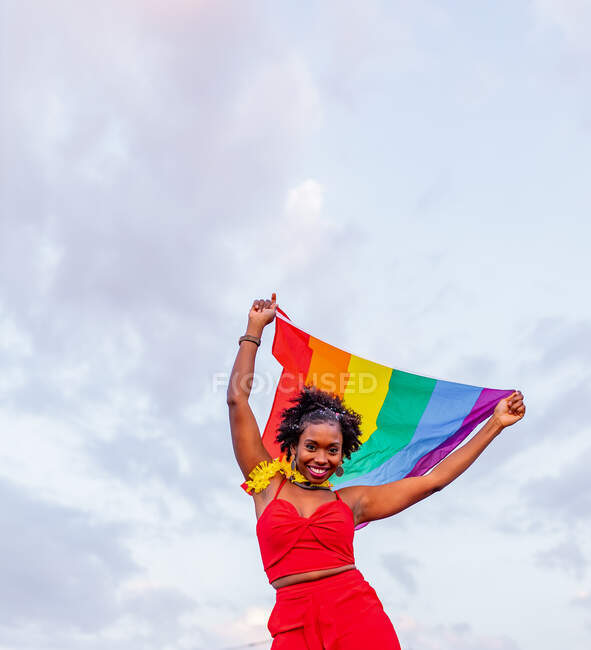 Desde abajo de la mujer afroamericana con estilo en el desgaste de moda izamiento de la bandera con adorno de arco iris mientras mira a la cámara en la carretera - foto de stock