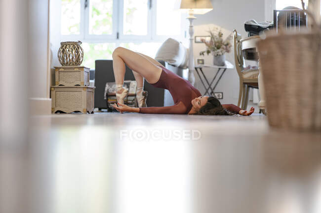 Vista lateral a nivel del suelo de la esbelta bailarina de ballet en traje de cuerpo y zapatos puntiagudos tumbados en el suelo en casa - foto de stock