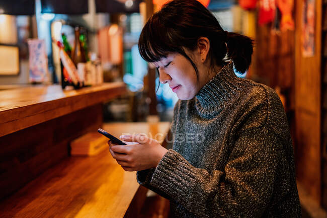 Vista lateral da senhora asiática satisfeita em camisola casual sorrindo ao usar o telefone móvel no balcão no bar ramen tradicional — Fotografia de Stock