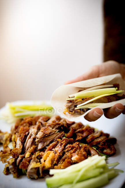 Hands of woman sitting at dinner table and holding Beijing duck piece on rice flat bread over ceramic plate — Stock Photo