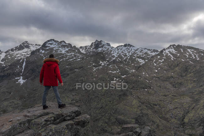 Vista posteriore dell'uomo irriconoscibile in capispalla in piedi sulla pietra e guardando la cresta innevata della Sierra de Gredos in una serata nuvolosa ad Avila, Spagna — Foto stock
