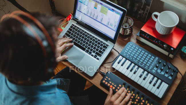 From above unrecognizable of focused young man in headphones working on synthesizer and laptop at table at home — Stock Photo