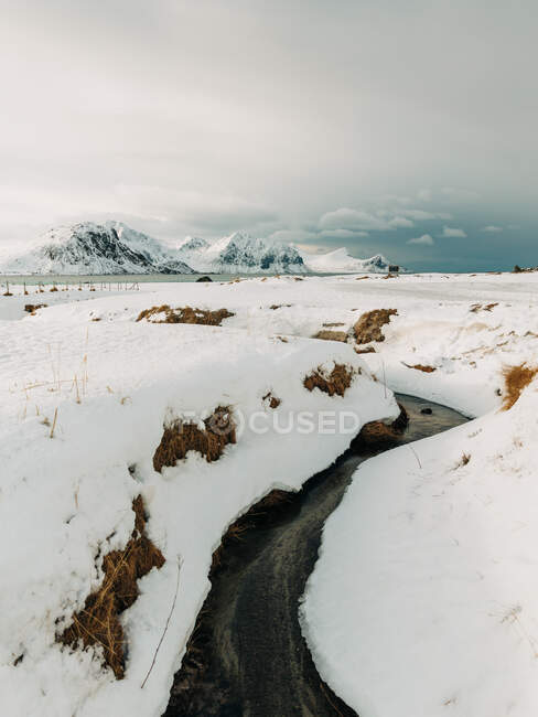 Narrow creek with cold water flowing under snow against mountains and overcast sky on Lofoten Islands, Norway — Stock Photo