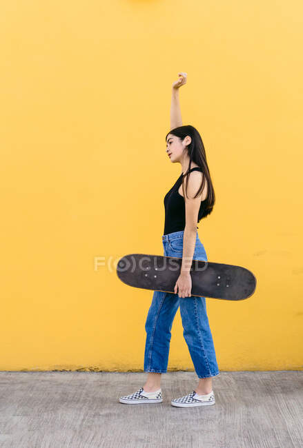 Vista lateral de la joven patinadora femenina con el brazo levantado y el monopatín de pie mirando hacia otro lado en la pasarela con colorida pared amarilla en el fondo durante el día - foto de stock