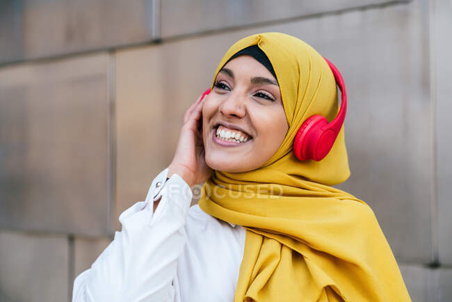 Allegro musulmana femminile in hijab ascoltare musica in cuffie su sfondo piastrelle in città — Foto stock