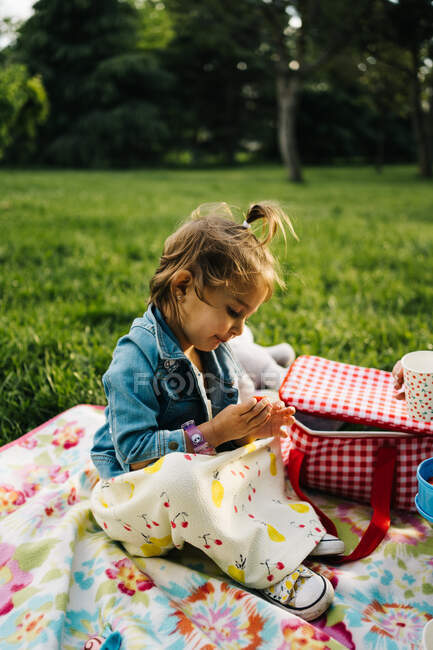 Vista lateral de linda niña en vestido y chaqueta de mezclilla jugando en manta de picnic colorido en el césped verde en el parque de verano - foto de stock