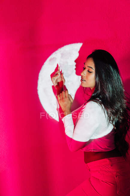 Vue latérale de la jeune femme hispanique debout avec les yeux fermés sous l'éclairage rose vif néon et projection de la lune — Photo de stock