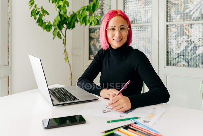 Diseñadora freelance femenina sonriente pensativa en atuendo casual y gafas mirando a la cámara mientras trabaja en la mesa con el portátil y dibuja bocetos de moda con lápices - foto de stock