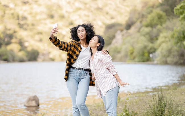 Content couple of multiracial lesbian females pouting lips and taking selfie on smartphone while hugging on shore of lake in forest — Stock Photo