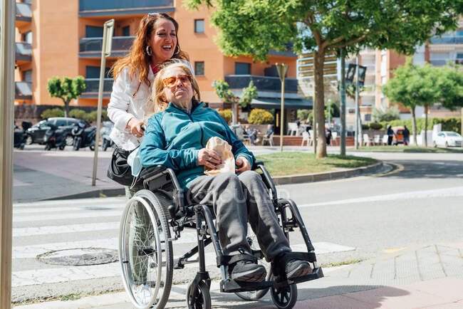 Erwachsene Frau schiebt Rollstuhl mit Seniorin und überquert Straße bei Sommerspaziergang in der Stadt — Stockfoto