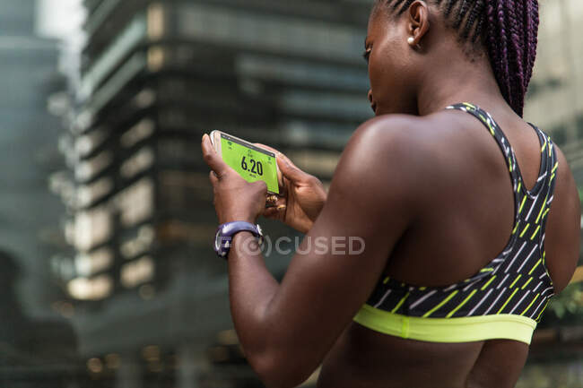 Невпізнавана афро-американська жінка, яка переглядає фітнес-додаток на смартфоні, стоячи на розмитому тлі вулиць міста під час тренування на відкритому повітрі. — стокове фото