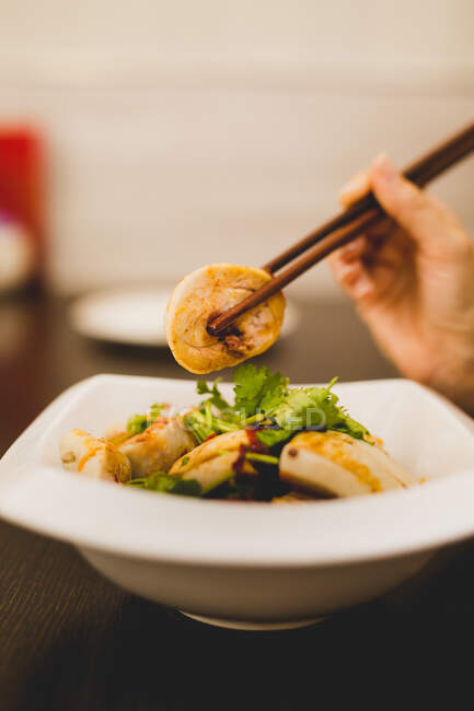 Hand of woman sitting at table and eating with chopsticks Chinese meat roll from white ceramic plate — Stock Photo